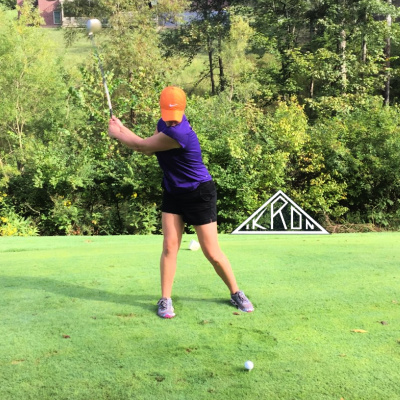 A woman in a purple shirt and orange hat has raised her golf club in preparation to hit the ball in front of her. In the backgroud are trees and the IKRON logo